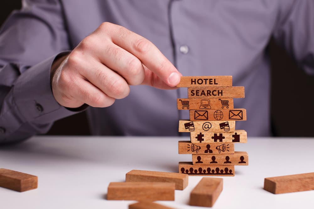 Hand, die Bausteine mit den Worten 'Hotel' und 'Search' sowie Online-Marketing-Symbolen aufstellt
