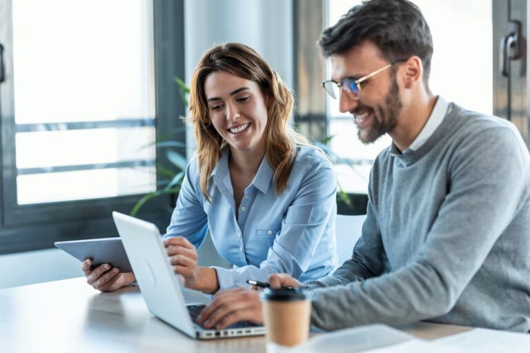 Zwei lächelnde Marketing-Profis arbeiten gemeinsam an einem Laptop und besprechen Strategien